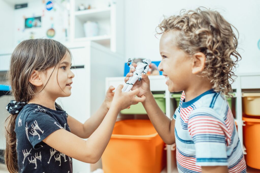 Two children enjoy playtime indoors, sharing a toy with joyful expressions.
